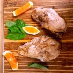 Top shot of two sous vide chicken breasts on wooden cutting board next to orange slices.
