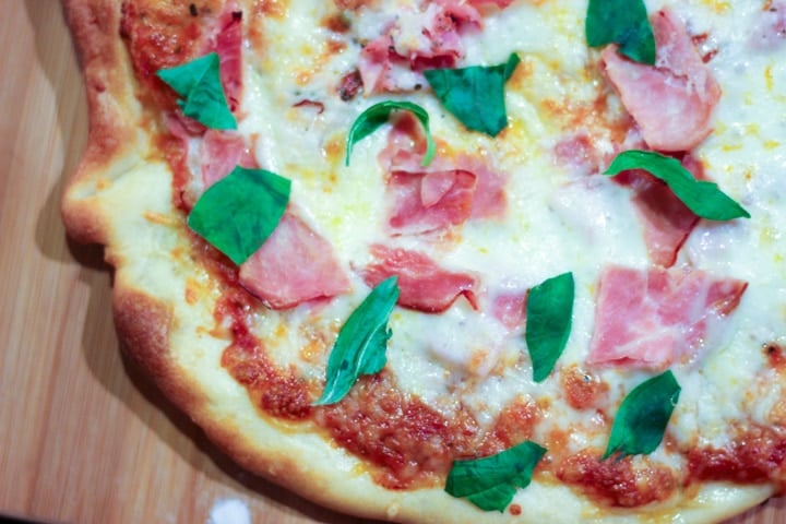 Chewy pizza crust on homemade pizza cooling on a cutting board topped with fresh basil ham and mozzarella