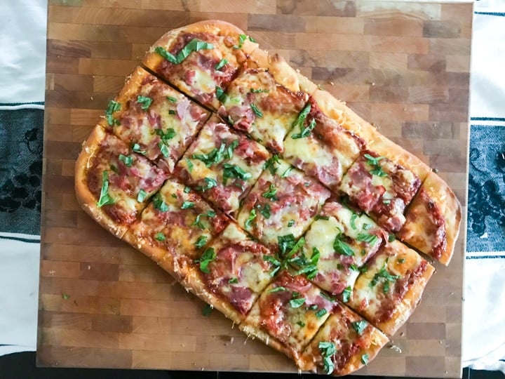 homemade pizza topped with ham cheese and basil shaped in a rectangle cooling on a butcher block