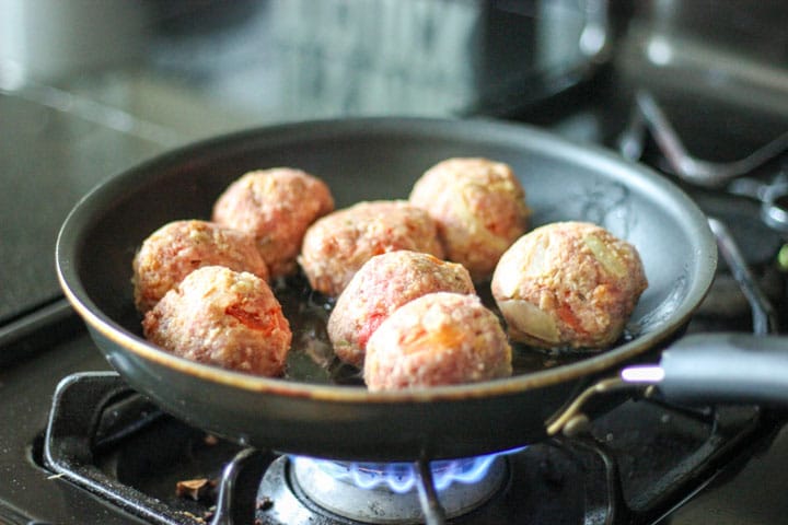 Raw meatballs cooking on the stove in black skillet.