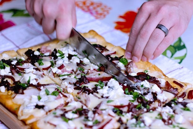 cutting pizza on a cutting board featuring pear and goat cheese flatbread topped with caramelized onions, ham and cheddar