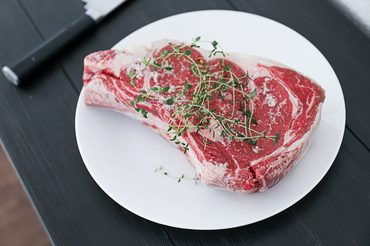 Top shot of uncooked prime rib steak garnished with thyme sprig on white plate