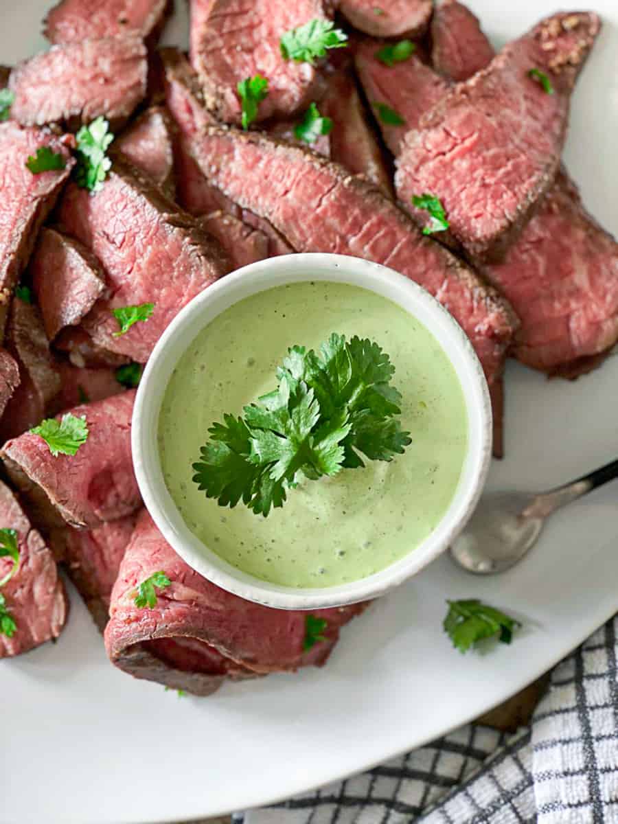 sous vide tri tip steak sliced on white plate next to small white bowl of green sauce