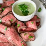 sliced steak on white plate next to small white bowl of green sauce