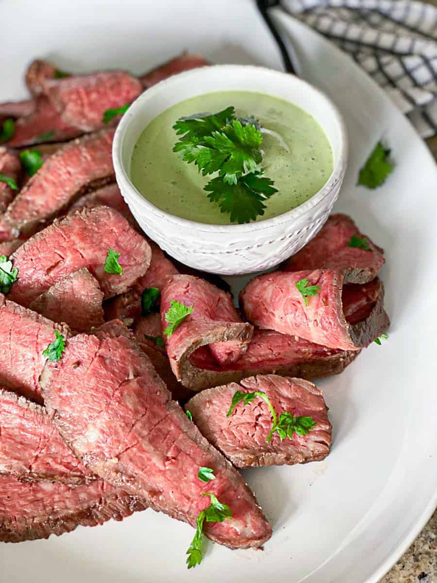 sliced steak on white plate next to small white bowl of green sauce