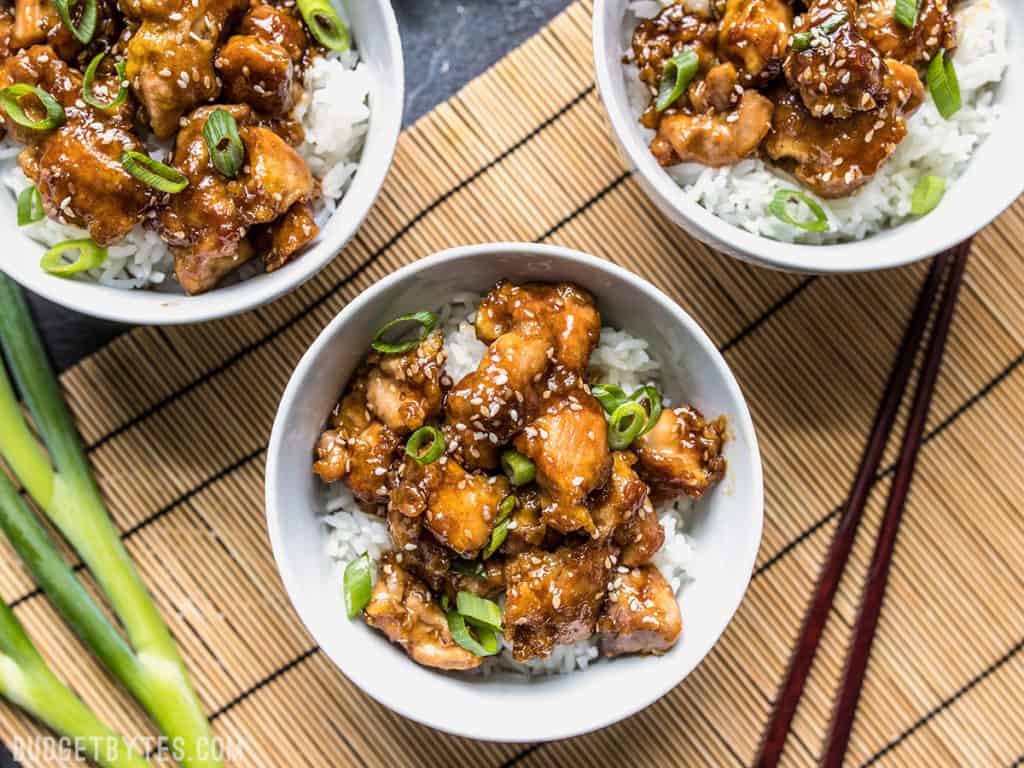 three sesame chicken with rice and green onion bowls placed on top of a bamboo mat with chopsticks