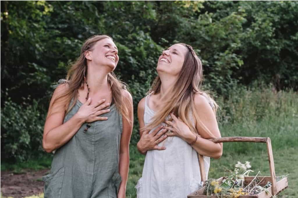 2 women are laughing while one woman is carrying a basket of foraged plants