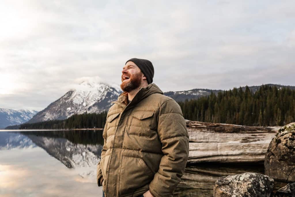 man with mouth open smiling wearing a hat and jacket standing in front of a lake and mountains