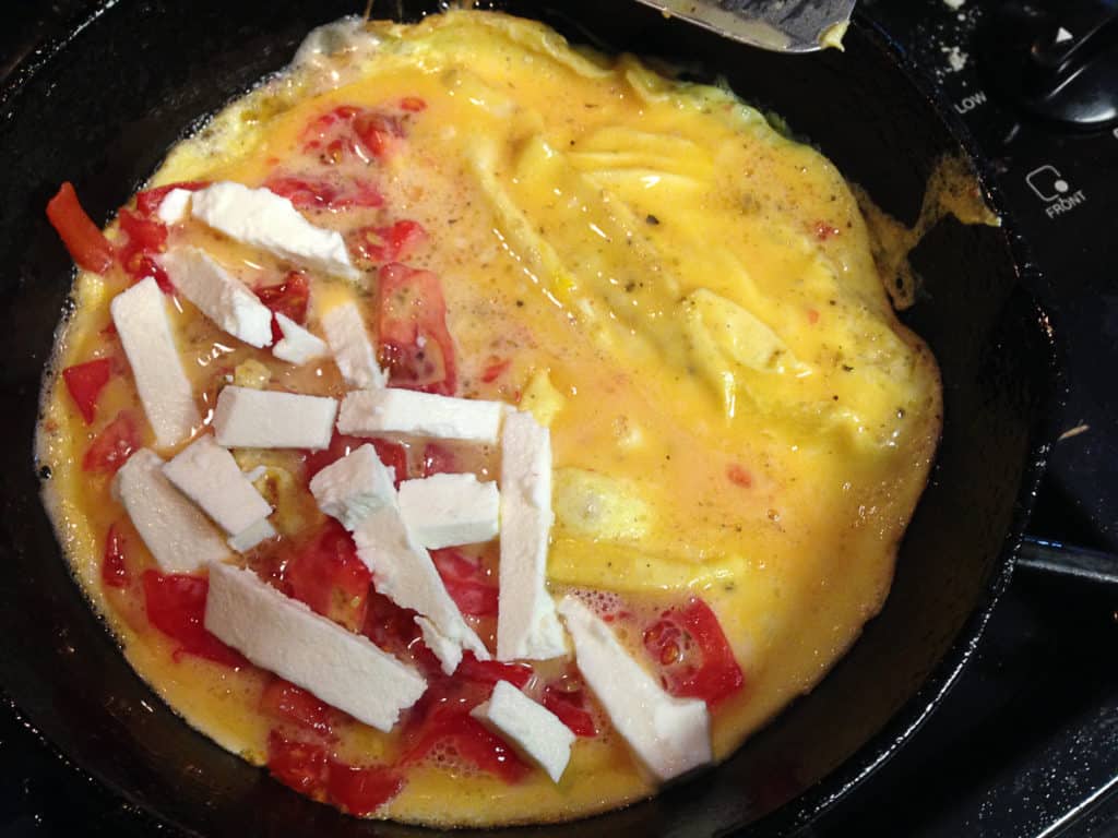 an omelet half cooked in a cast iron skillet with feta cheese strips and tomatoes on the left side of the omelet