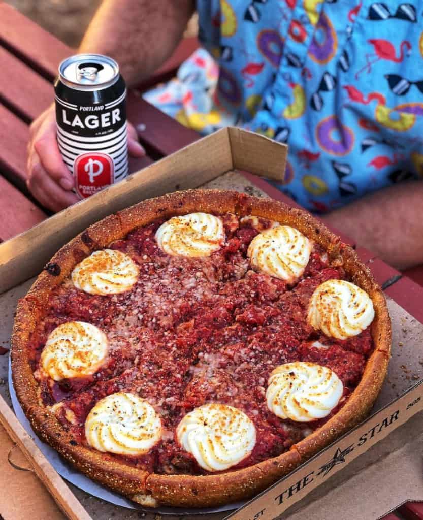deep dish pizza in box with a man holding a tall can of beer next to it