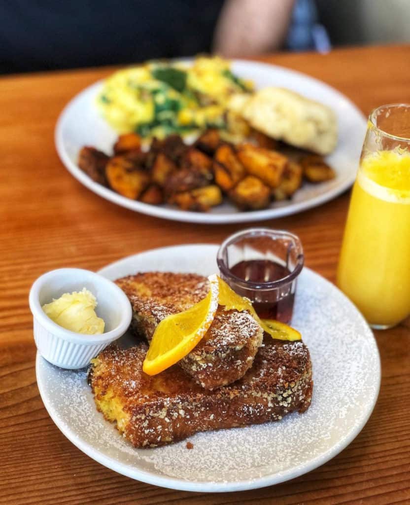 french toast with a small ramekin of butter and container of maple syrup on a plate, dusted with powdered sugar and topped with a spiraled slice of orange, a mimosa in a stemless glass next to the plate