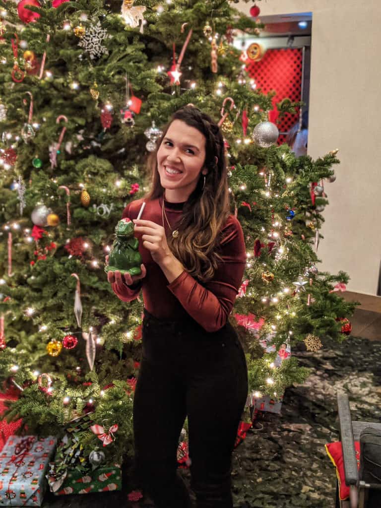 woman smiling standing in front of a christmas treen with a cup shaped like a trex in a santa hat