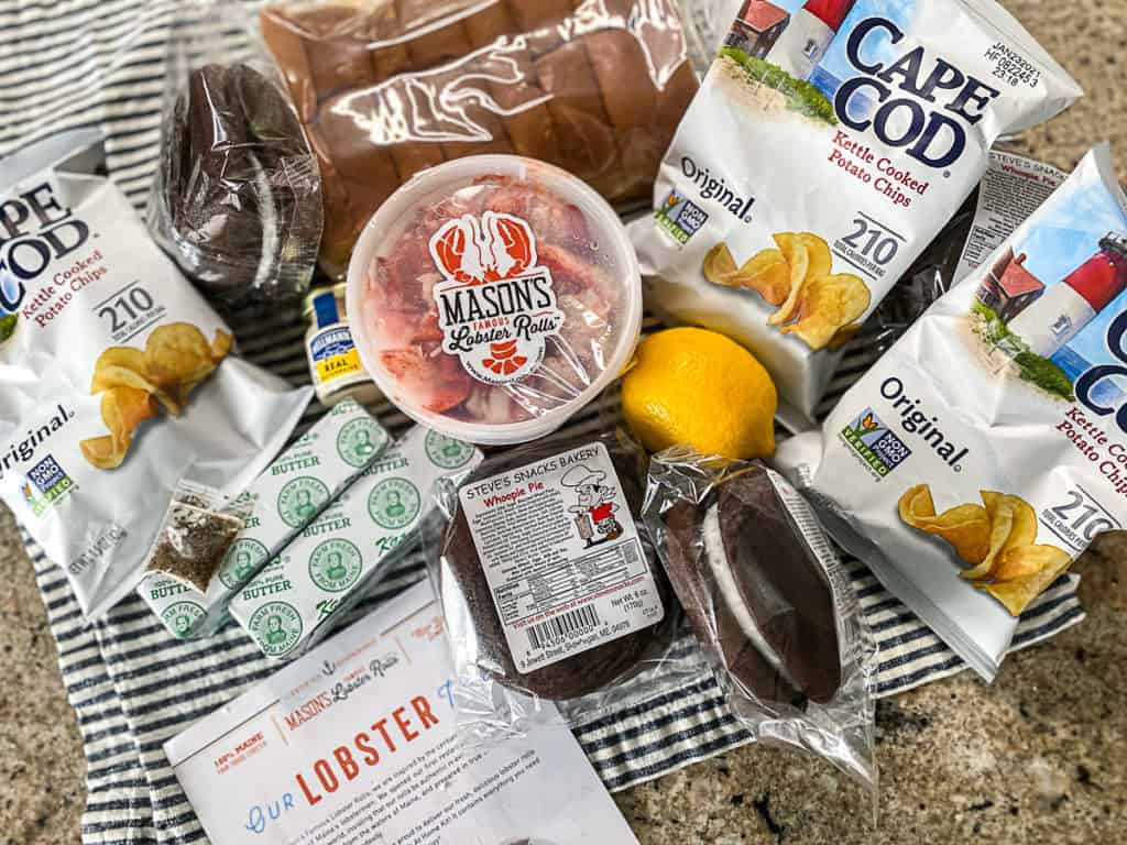Lobster roll kit with three bags of cape code potato chips, three whoopie pies individually wrapped, a lemon, butter, mayo, container of lobster meat, and buns in a bag