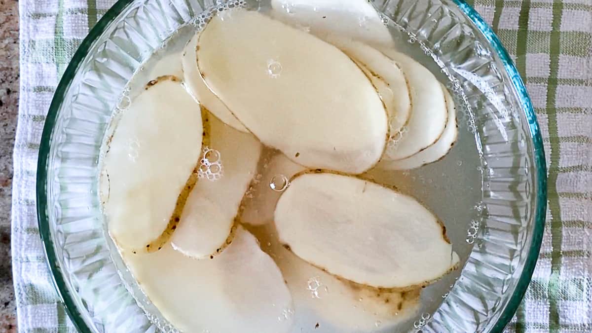 Thinly sliced potatoes soaking in water in glass mixing bowl.