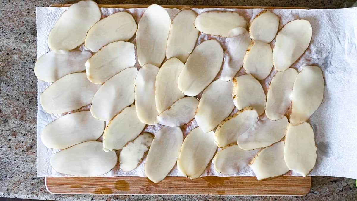 Thinly sliced potatoes arranged in rows on paper towel on wooden chopping block.