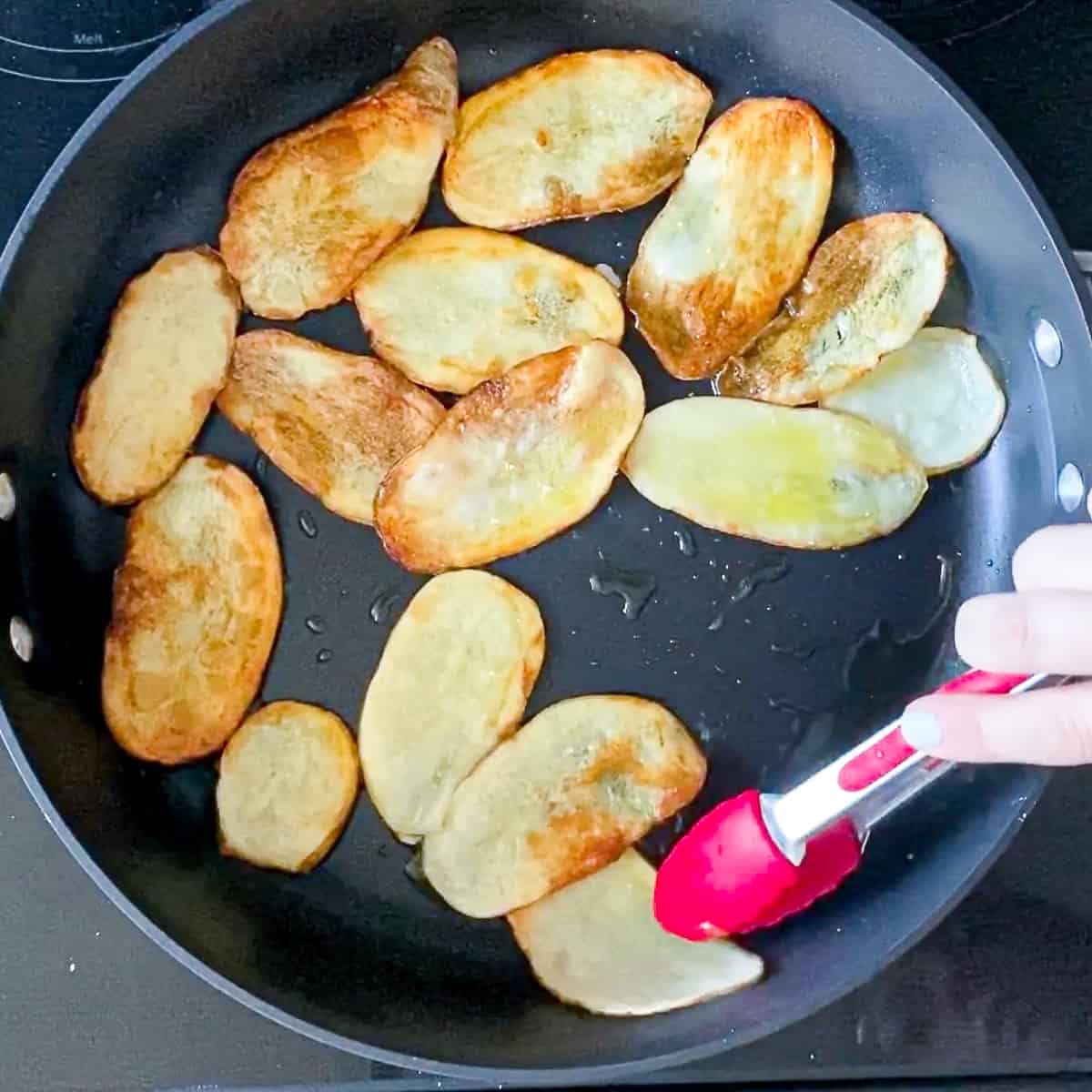 Tongs moving thinly sliced potatoes around on skillet.