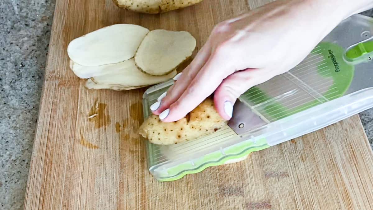 Potato being sliced with green mandoline on wooden cutting board.