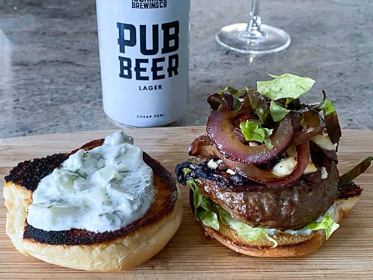 Fully dressed sous vide lamb burger with onions, lettuce, crumbled cheese and tzatziki sauce on wooden cutting board.