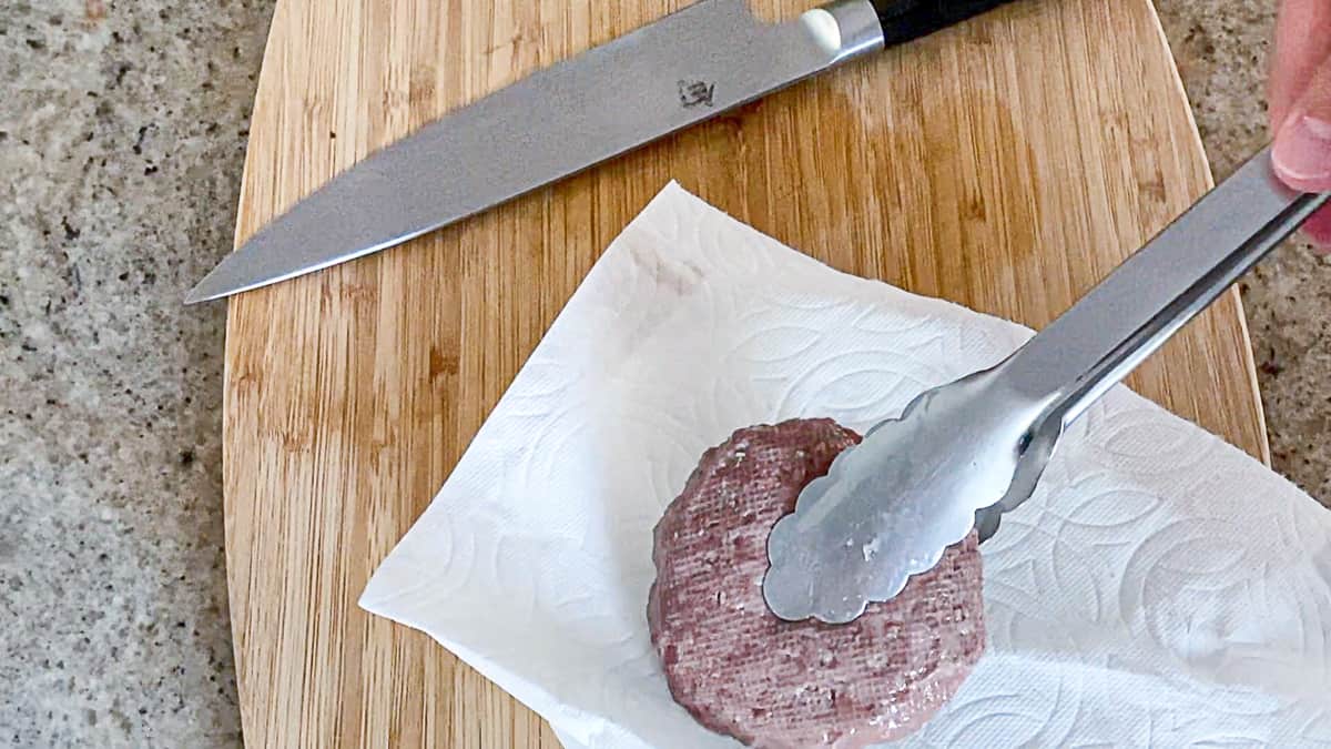Tongs holding lamb burger patty over paper towel after sous vide bath.