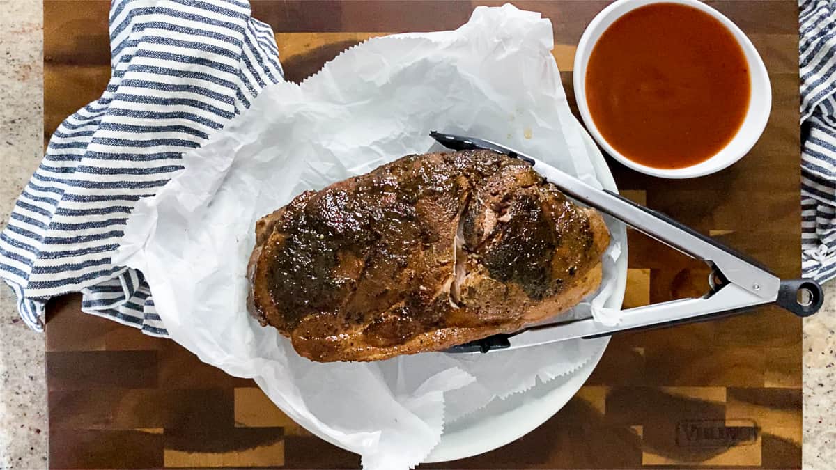 Cooked pork shoulder next to tongs and cup of BBQ sauce.
