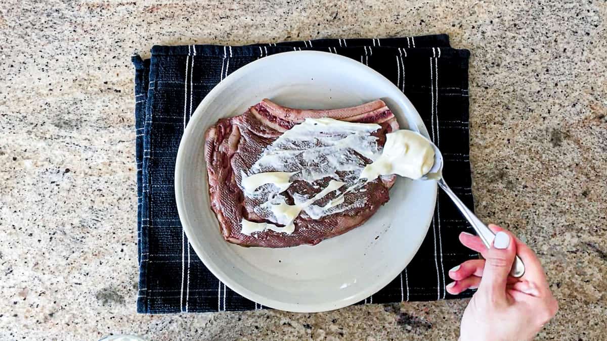 Spreading mayonnaise with spoon on top of steak, sitting on white plate and tea towel on countertop.