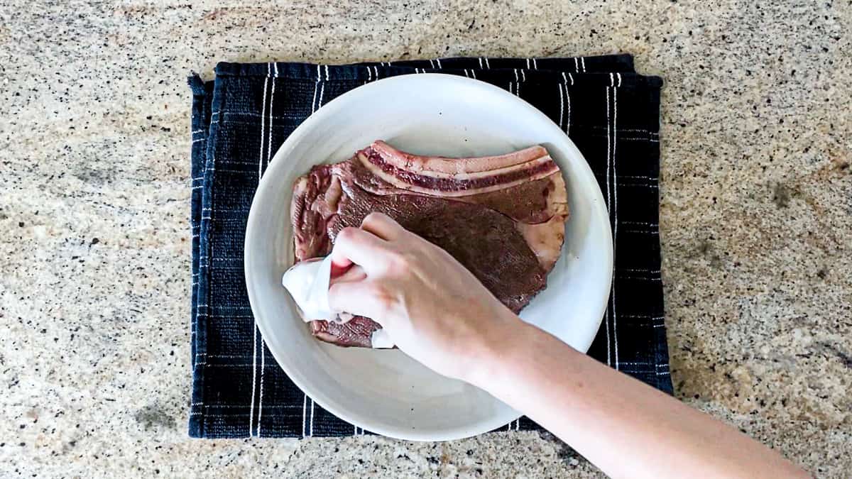 Patting dry a steak after sous vide bath, steak is sitting on white plate and tea towel on countertop.