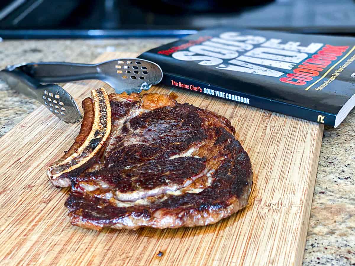 Cooked sous vide ribeye steak on wooden cutting board next to tongs and cookbook.