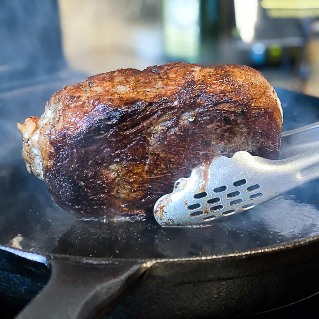 Closeup of tongs searing ribeye steak in a cast iron skillet.