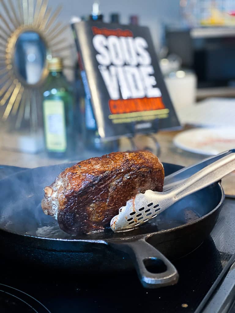 Tongs holding sous vide ribeye steak with the home chef's sous vide cookbook in background