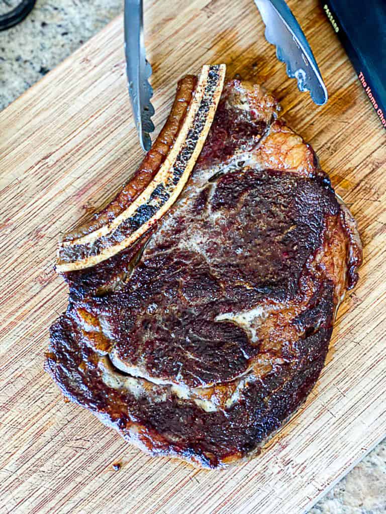 Top shot of bone in ribeye steak with tongs on a cutting board.