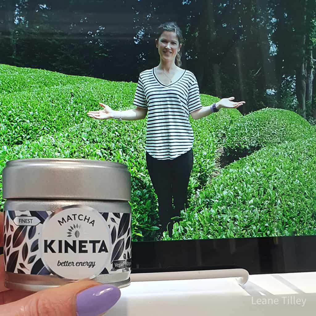 close up of hand holding a contatiner of matcha in front of a picture of a woman standing in greenery