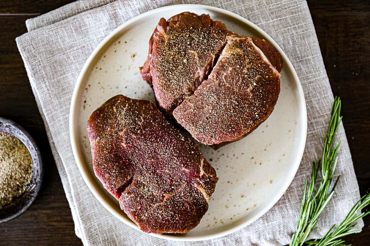 Seasoned filet mignon steaks for air frying with a dry rub bowl and fresh herbs on the side.