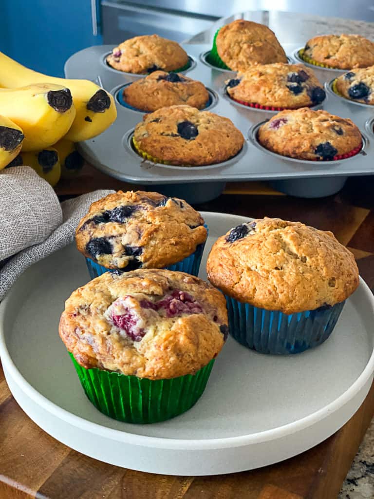 Side shot of fresh banana blueberry muffins on a plate with a muffin tin in the background.