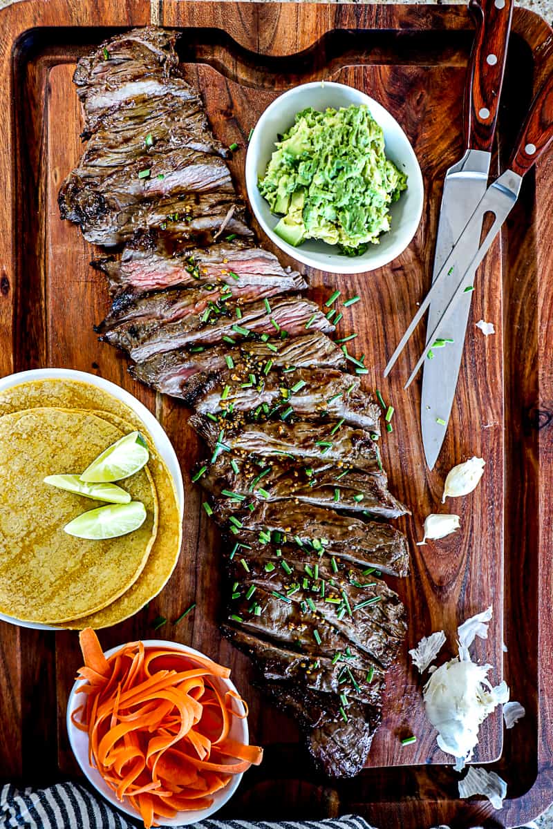 Top down shot of grilled skirt steak recipe on a large party platter cutting board with taco shells, lime, and guacamole.