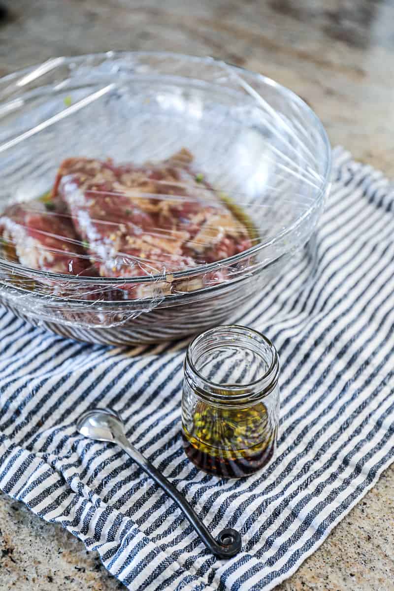 Side shot of skirt steak marinading in a large bowl wrapped in plastic wrap with a side marinade in a jar and a spoon on a counter.