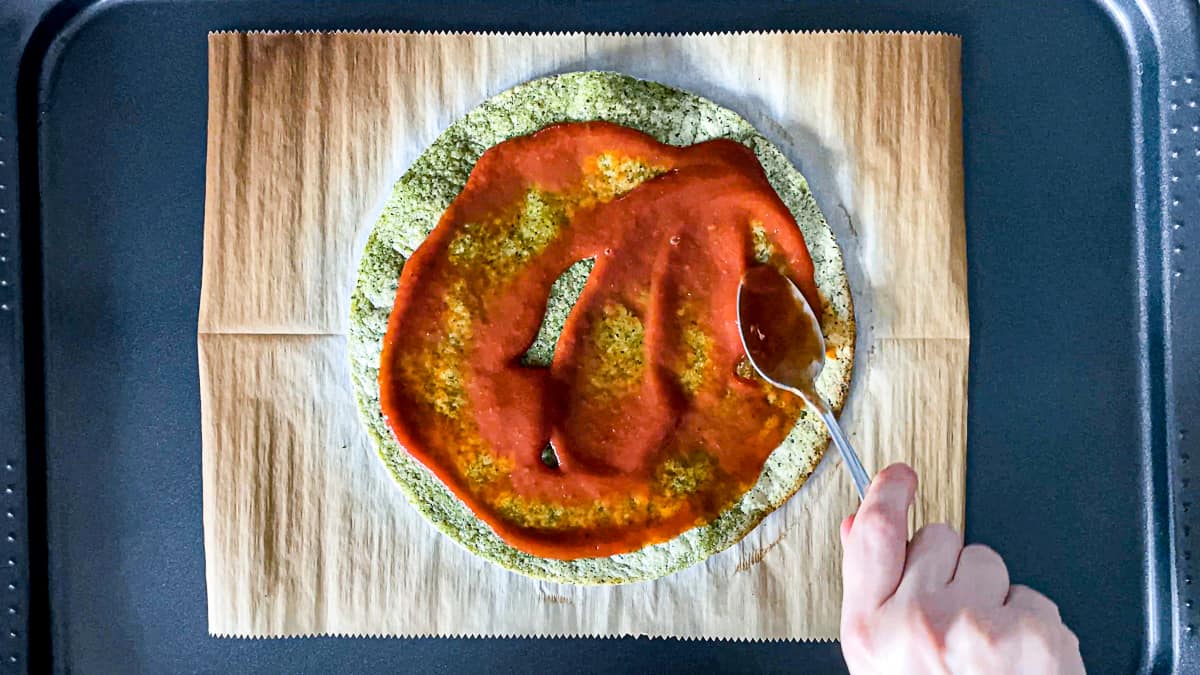 Top down shot of hand topping parbaked broccoli pizza crust with pizza sauce.