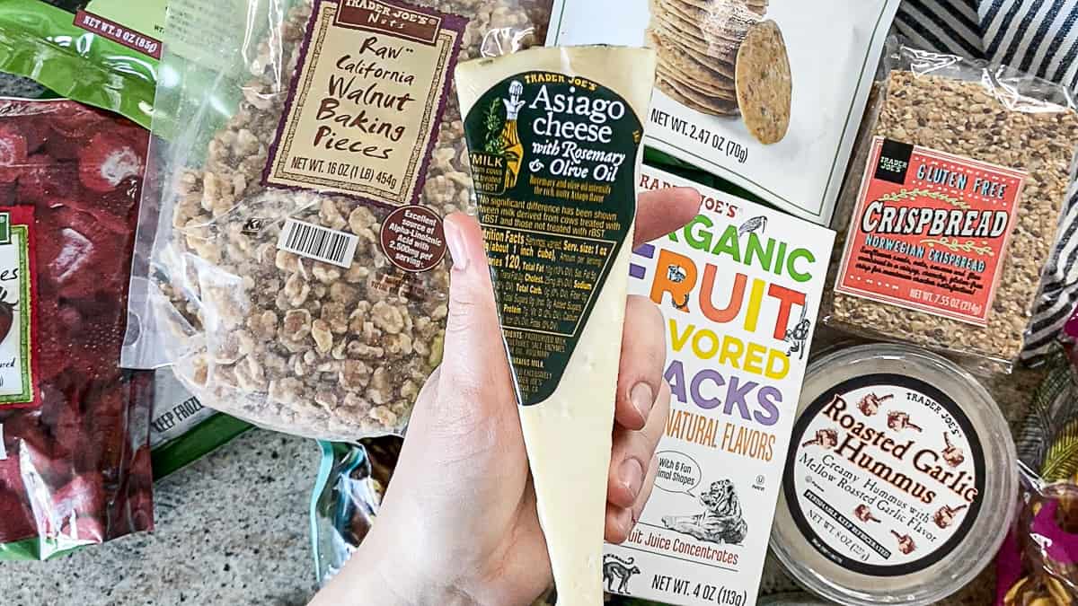 Top down shot of Rosemary Asiago Cheese from Trader Joe’s with other shopping haul items including crispbread, hummus, walnuts, dried strawberries.