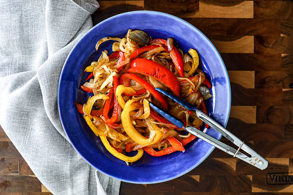 Top down shot serving peppers and onions in a side dish with tongs for steak and chicken and shrimp.
