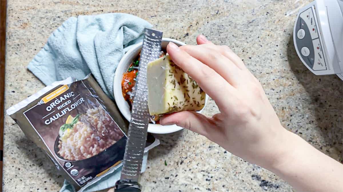 Top down shot of Trader Joe's cheese being grated by hand into a bowl of meatballs with a side bag of cauliflower rice.