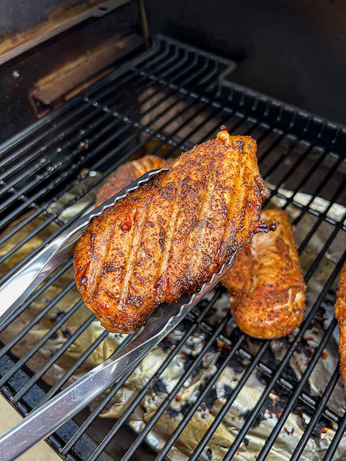 Tongs holding smoked chicken breasts over Traeger pellet grill