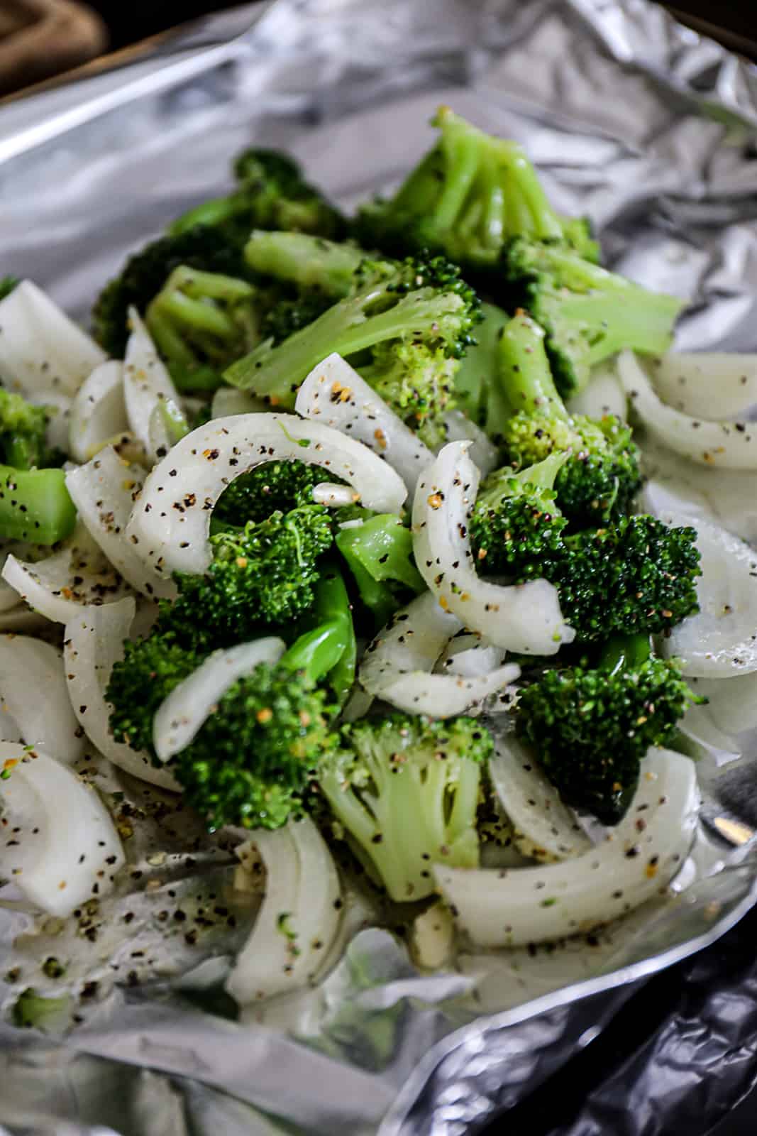 Closeup of seasoning broccoli and onions for grilling on foil