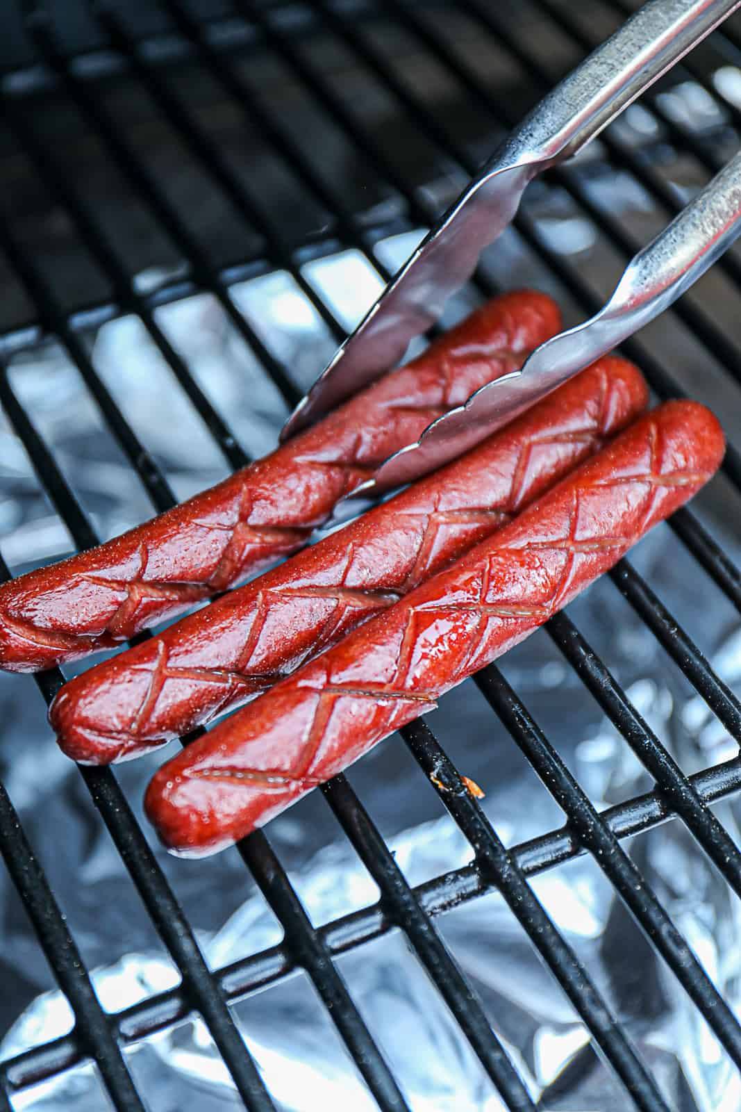 Tongs Holding Scored Smoked Hot Dogs On Traeger Grills 