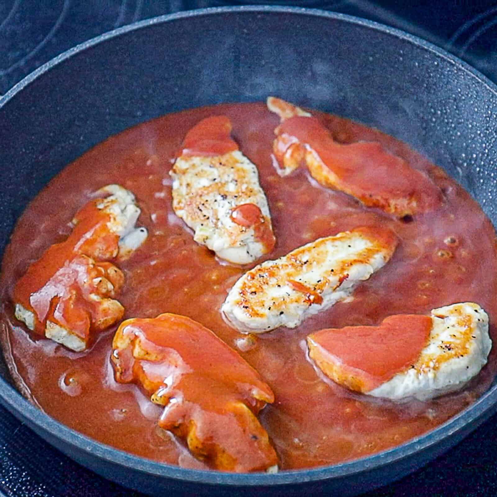 Chicken tenderloins flipped and simmering in enchilada sauce in a pan
