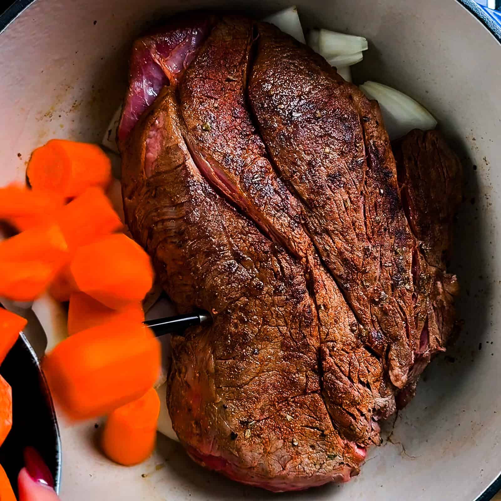 Dutch oven on the stove with seared chuck roast, onions, and diced carrots